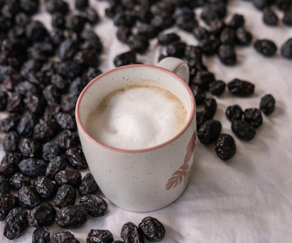 A mug of cappuccino with foam surrounded by dried black olives on a white linen table.